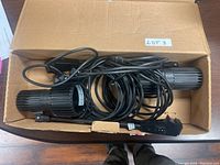 Two black Skandalight exhibition stand lights inside an open cardboard box with cords coiled around them and a small LOT3 label attached.