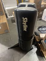 Exterior view of the black cylindrical Skyline transport case standing upright on the floor among boxes.