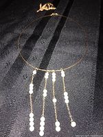 Gold-colored wire choker necklace with five hanging chains adorned with small white faux pearls, laid on black fabric.