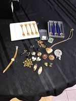 Full view of the lot showing jewelry items including brooches, earrings, a necklace, and collectible spoons laid out on a black tablecloth.