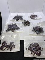 Top-down view of 7 brooches arranged on white paper, each in a clear plastic bag except one in center