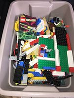 Top view of container showing assorted LEGO pieces including LEGO System booklet, colorful bricks, wheels, large baseplates, and structural parts inside a white Rubbermaid Roughneck bin.