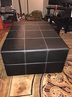Large black faux leather ottoman with white grid stitching showing wear and damage on surface, situated on patterned carpet in a home living room environment.