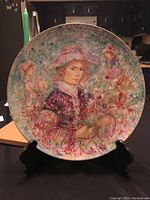 Full view of the decorative plate on a stand showing the central image of a girl with a large hat and floral bouquet in soft pastel colors.