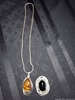Photo showing the silver necklace with amber pendant and silver brooch with black stone on a black fabric background.