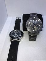 Two watches displayed side by side on a white surface. One is a silver stainless steel Fossil chronograph watch, the other is a black dial watch with a black rubber strap.