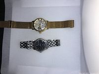 Two men's wristwatches laid flat: one gold-tone skeleton dial watch with mesh bracelet, one silver-tone Guess watch with black face and metal link bracelet.