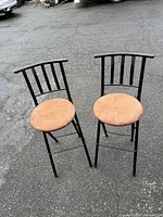 Two black metal frame bar stools with brown round cushioned seats and vertical bar backrests placed on asphalt ground outdoors.
