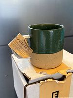 Photo of the ceramic mug on top of its original box, showing the handle and the tag attached indicating it's from United by Blue Ceramic Collection.