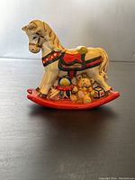 Front view of ceramic rocking horse coin bank showcasing the colorful saddle, horse mane, and toy decorations on the rocking base including teddy bear and globe.