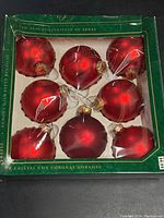 Top view of green box containing nine red glass Christmas ball ornaments with gold crown tops sealed with plastic.