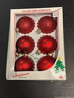 Full view of the box containing six red glass Christmas ball ornaments, three glittery and three smooth.