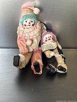 Full view of both clown dolls sitting against a metallic background, showing fabric costumes and painted faces.