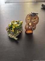 Photo showing green frog figurine sitting on a leaf-shaped base next to brown owl figurine.
