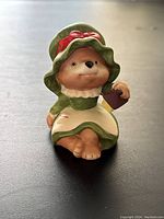 Front view of ceramic bear figurine in green bonnet and dress with red bow and white apron holding small gift box.