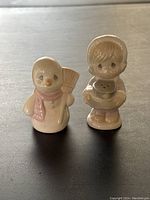 Two ceramic snowman-themed salt and pepper shakers, one snowman with pink scarf and broom, other child holding a snowman figurine.