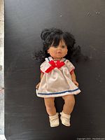 Full view of the vintage 14 inch BSB Baby So Beautiful doll lying on a black surface, showing the full body in the white dress with red and blue trim and white shoes.