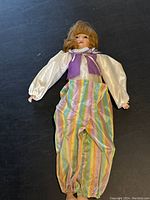 Full front view of vintage clown doll lying on black surface, showing colorful striped pants, purple vest, and white blouse with puffy sleeves.