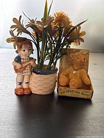 Set of three vintage figurines placed on a surface including a boy figurine with floral flowers and a teddy bear in a box