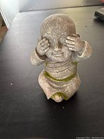 Front view of a small grey Zen monk statue with moss detailing covering eyes with hands