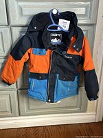 Full view of the boys winter jacket showing multi-color design in orange, blue, and black with front zipper and button pockets.