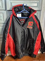 Front image of Calgary Flames men's jacket XXL in black with red and white accents on sleeves and sides, hanging on blue hanger.