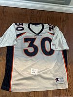 Front view of vintage white Denver Broncos football jersey with number 30 and orange and navy trim on wooden floor.