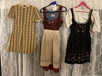 Photo showing three dresses hanging: yellow 1970s mini dress, brown dirndl dress with apron, and black floral dirndl dress with blouse.