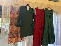 Four dresses hanging on curtain rod: patterned sleeveless, dark green long sleeve, red sleeveless midi, green sleeveless midi.