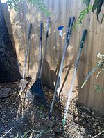 Photo showing six gardening tools leaning against a wooden fence with natural ground and leaves around them.