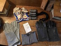 Two pairs of leather gloves, one pair of beige and blue fabric gloves and two striped cloth wristlets on wooden surface.