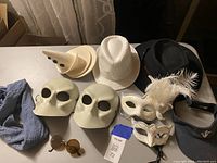 Photo showing two white Bauta masks, two cream cone-shaped hats with black buttons, white woven cowboy-style hat with feather, black fedora hat, grey scarf, round sunglasses, and white decorative masquerade masks.