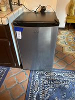 Closed view of stainless steel Whirlpool mini fridge with black top and handle showing front and part of surrounding room.