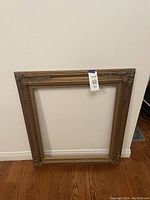 Dark gold rectangular ornate frame leaning against wall on hardwood floor, tag with lot number attached.