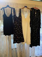 Photo showing four dresses hung on hangers against a light curtain. Colors include black, dark floral, white, and black with celestial print.