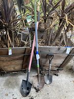 Three shovels standing upright against wooden planter background, showing size and blade shapes.