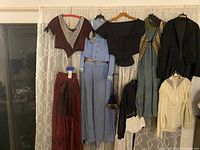 View of seven Victorian costumes hanging against curtain: red antebellum outfit, blue walking suit, black bodice, blue-green dress, men's black tail coat, white lacy blouse, and black cropped jacket with undershirt.