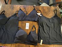 Four black women's exercise tops laid on a wooden surface, showing different styles: two bra tops and two tank tops with thin adjustable straps.
