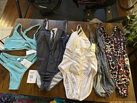 Five swimsuits laid out on a table including aqua two-piece, dark gray business suit-style one-piece, white ruched one-piece, and two patterned one-piece swimsuits.