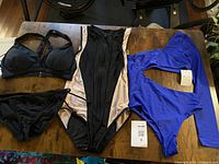 Photo of three women's swimsuits laid out on a wooden table including a black bikini set, beige and black one-piece with zipper, and blue two-piece with long sleeve top, all size large.