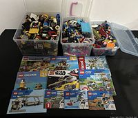 Three large clear plastic bins filled with assorted Lego bricks and various original instruction manuals laid out in front on a black surface.