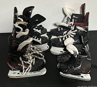 Four pairs of black ice hockey skates with white laces and blade holders, various sizes and styles placed side by side on a black surface.
