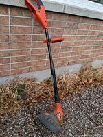 Side view of an orange and black Black and Decker weed trimmer standing upright against a brick wall outside. Shows adjustable handle grip and protective guard at cutting head.