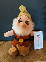 Front view of Doc plush toy wearing brown outfit, yellow hat, and white beard with Disney Store tag visible.