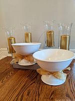Four gold candles in clear glass hurricane holders and two white ceramic pedestal bowls with gold bows on a wooden table.