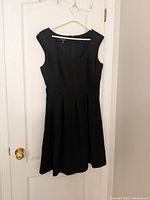 Full view of the black Nine West sleeveless dress with pleated skirt hanging on a white hanger on a door.
