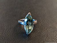 Front facing view of handmade silver ring with marquise-shaped greenish-blue semi-precious stone on black textured background.