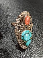 Close-up of silver ring showing teardrop turquoise and oval coral set in ornate silver band with scroll and floral patterns.