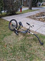 Full side view of the black SuperCycle ride-along bike trailer laying on the grass showing the frame, saddle, pedals, and attached tow arm.