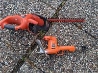 Two Black & Decker hedge trimmers placed on a pebble ground surface, one larger with a long blade and one smaller with short blade and orange body.